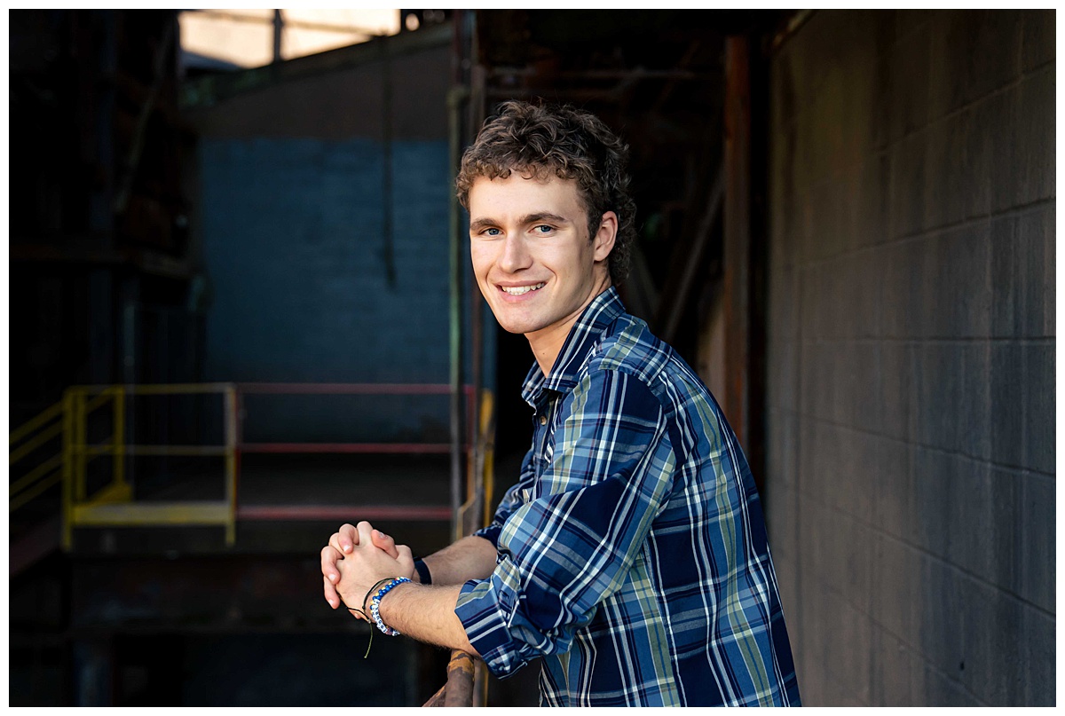 A young man poses for a portrait along a railing in an industrial setting.
